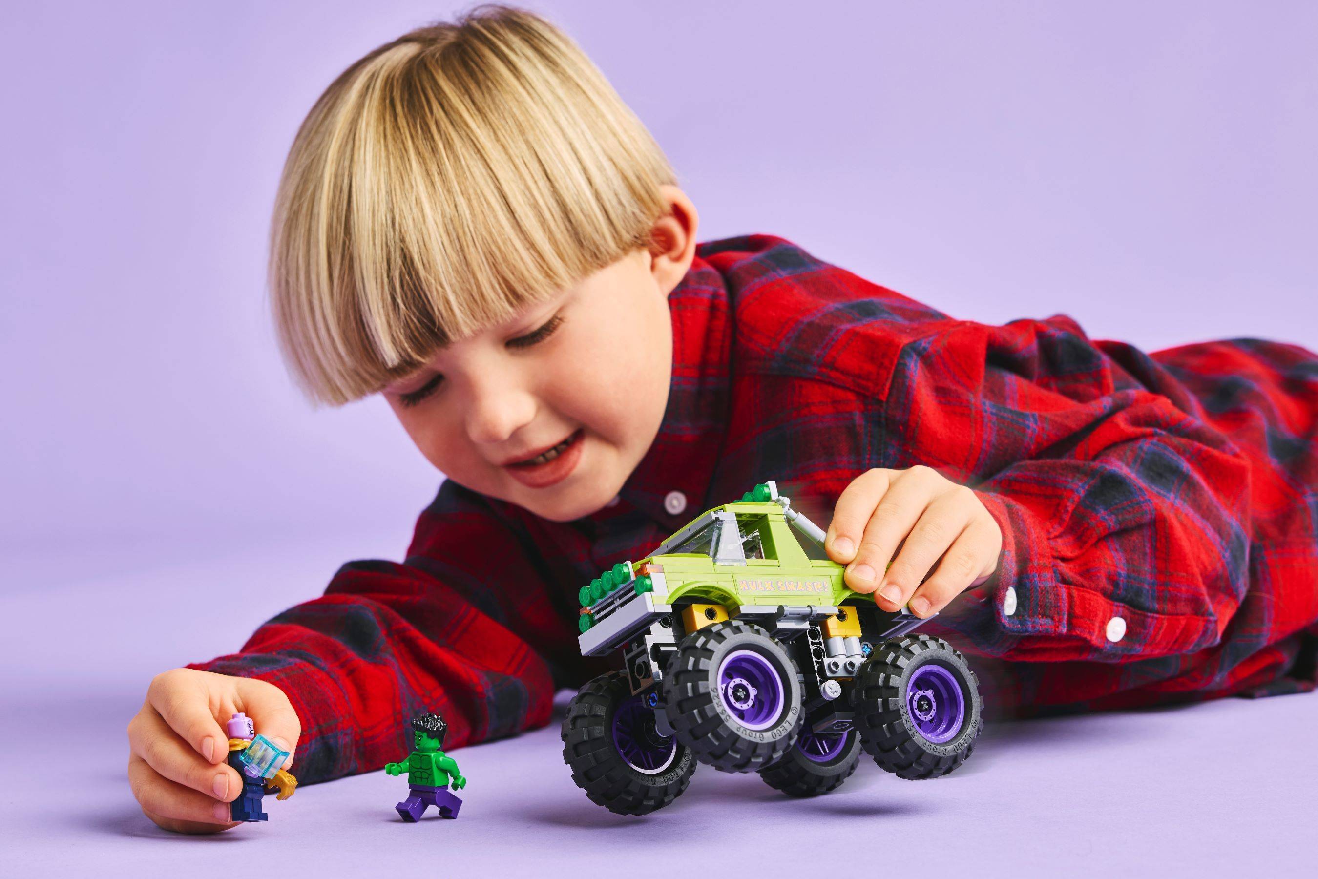Kleines Kind in einem roten Karohemd spielt mit Spielfiguren und einem grünen Monstertraktor auf violettem Hintergrund.