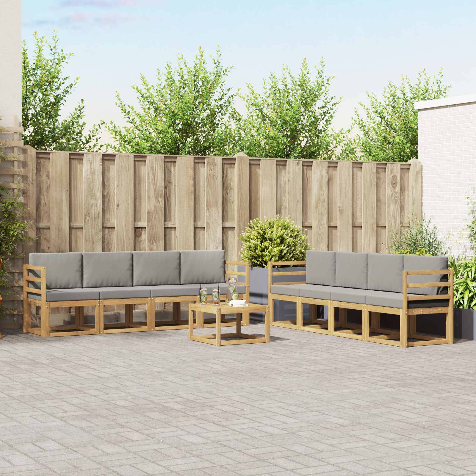 Außenbereich einer Terrasse mit zwei grauen, gepolsterten Holzsofas, einem kleinen Couchtisch und Topfpflanzen vor einem Holzzaun.