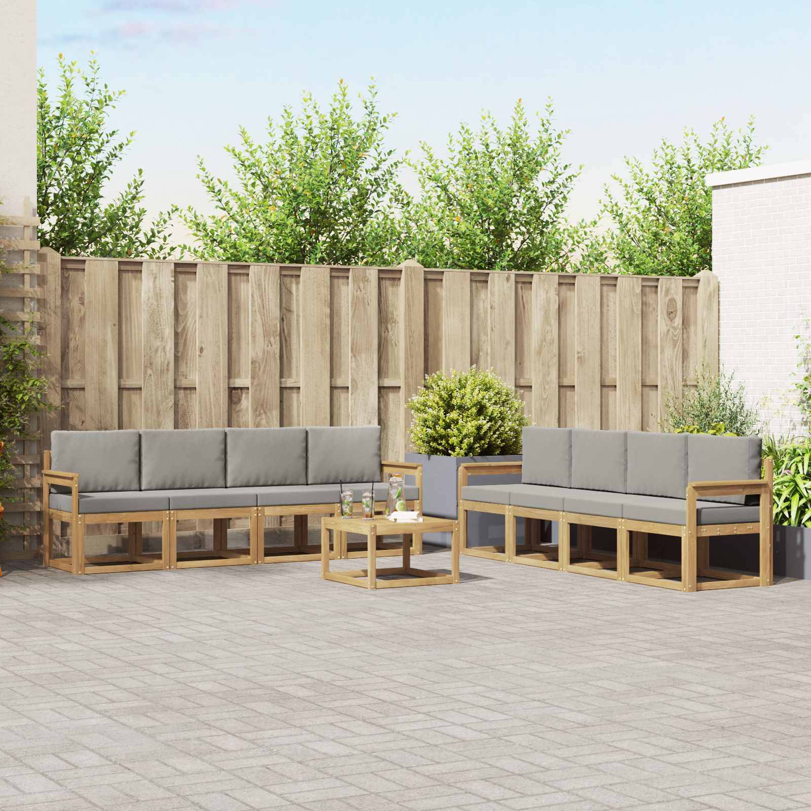 Outdoor-Terrassensitzbereich mit zwei graugepolsterten Holzsofas, einem kleinen Couchtisch, Topfpflanzen und einem Holz-Sichtschutzzaun.