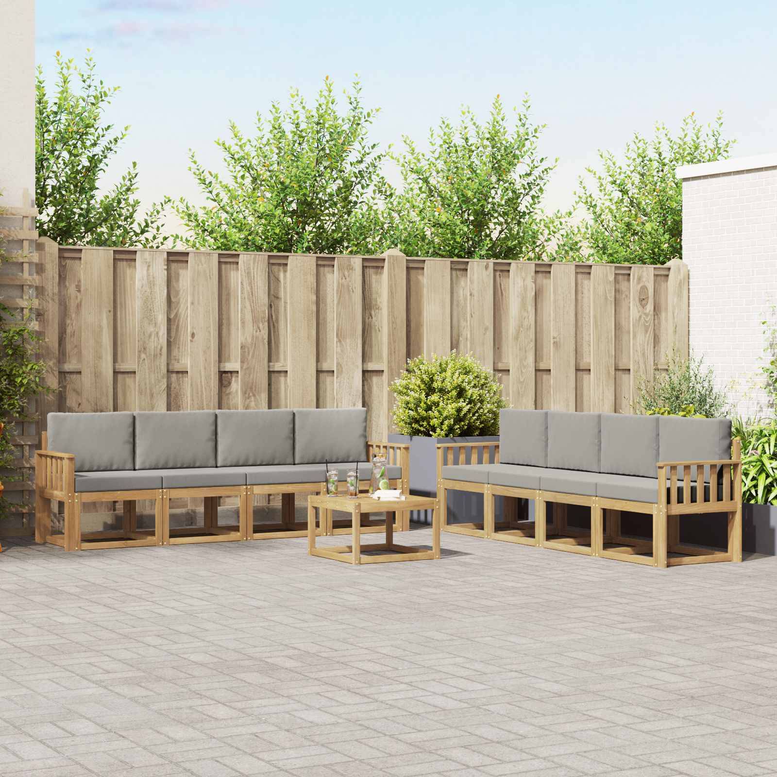 Sitzbereich auf der Terrasse mit zwei grau gepolsterten Holzsofas, die sich einem kleinen Couchtisch gegenüberstehen, vor einem hohen Holzzaun und Grünflächen.