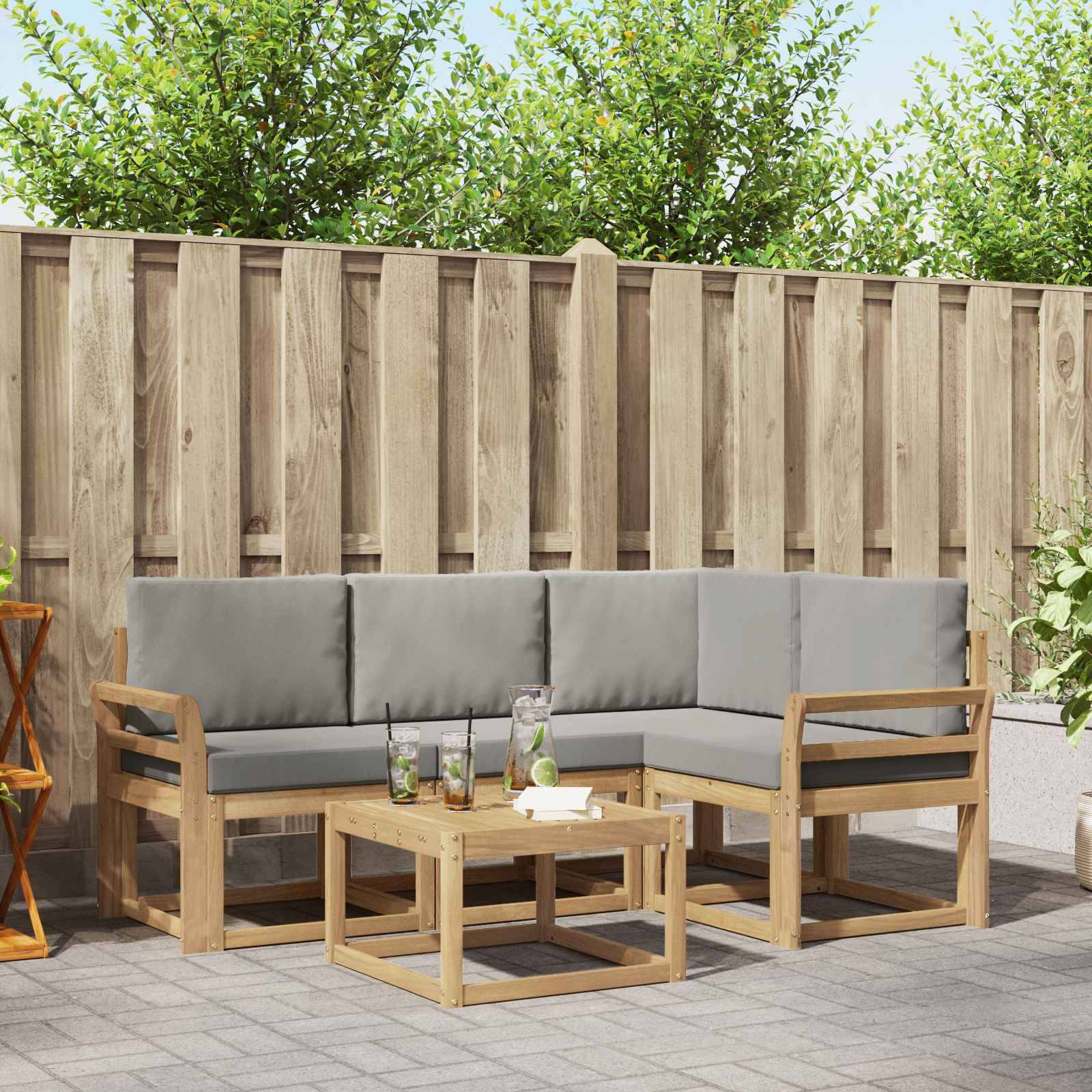 Außenbereich einer Terrasse mit grauem Ecksofa und hölzernem Couchtisch vor einem hölzernen Sichtschutzzaun und Grünflächen.