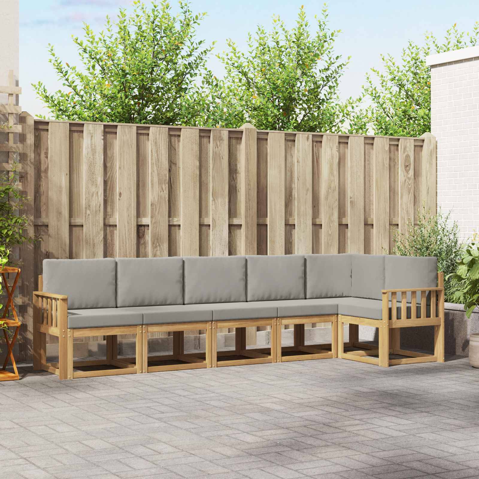 Outdoor-Ecksofa in L-Form mit grauen Kissen und Holzgestell auf einer gepflasterten Terrasse vor einem Holzzaun.