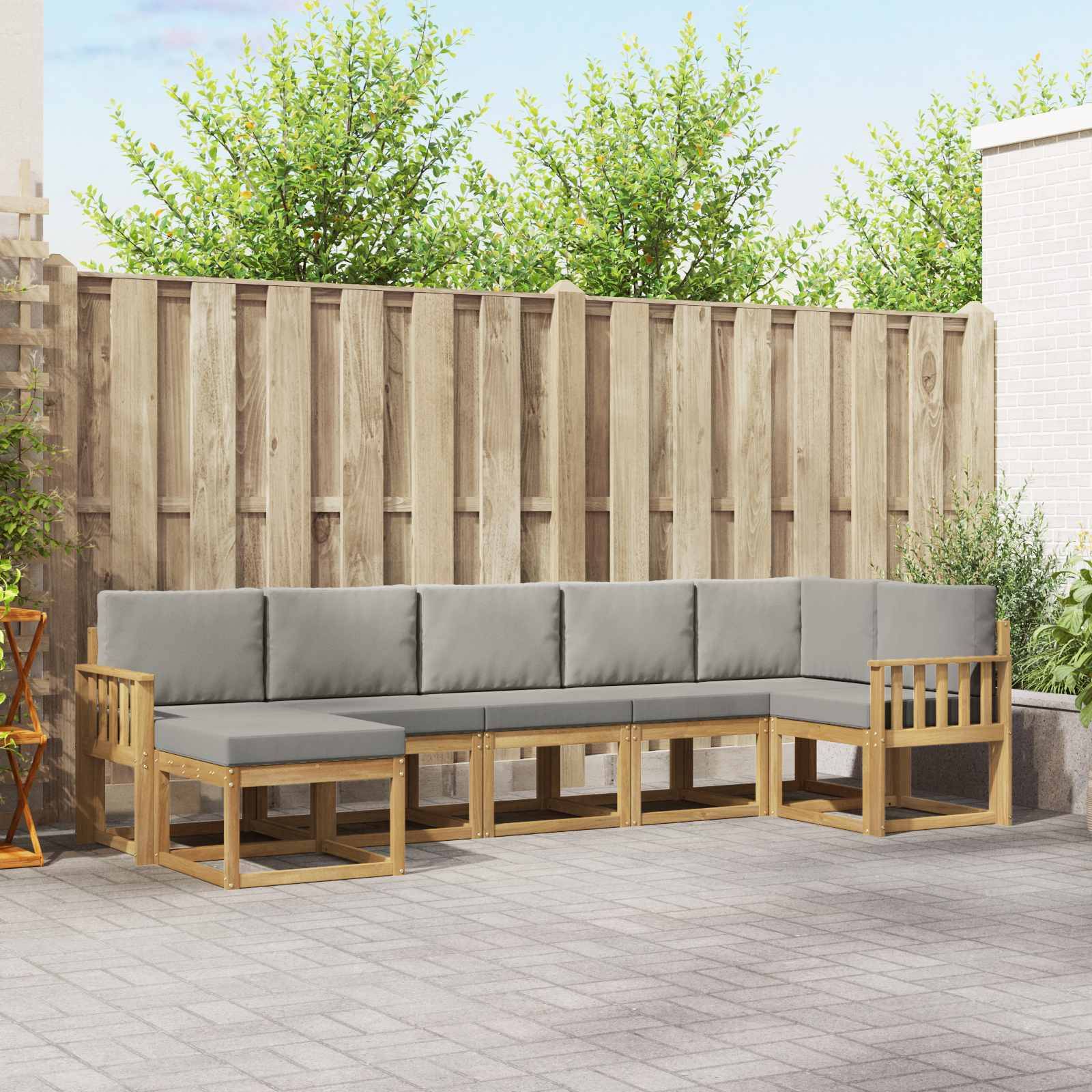 Außenbereich mit Sitzecke aus L-förmigem Holzsofa und grauen Kissen auf einer gepflasterten Terrasse, mit Holzzaun und Grünanlagen im Hintergrund.