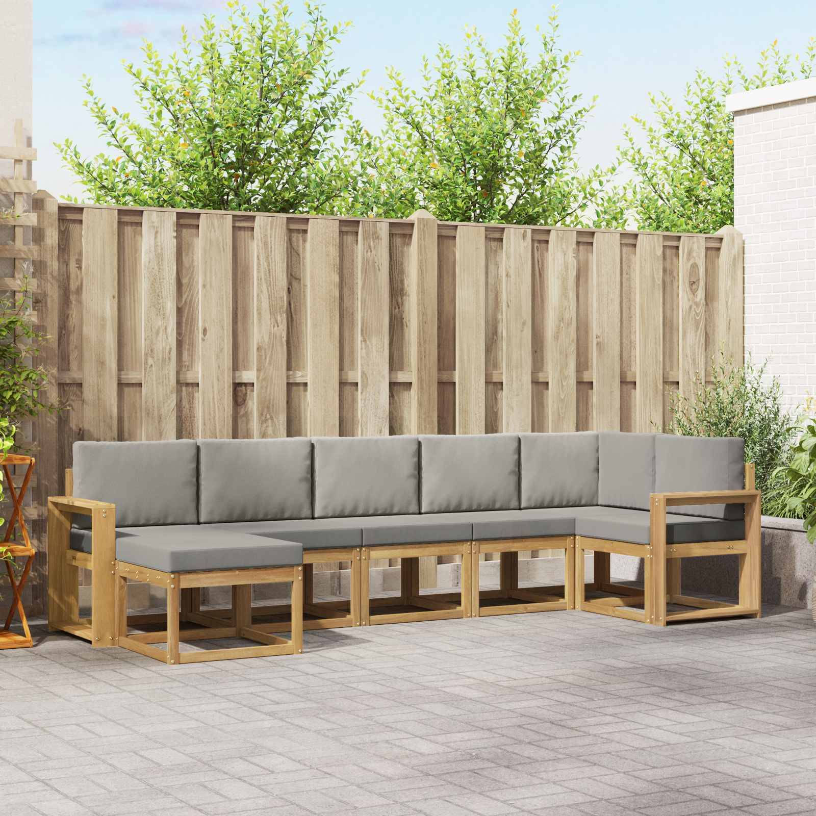 Outdoor-Ecksofa mit grauen Kissen und hellem Holzrahmen auf einer gepflasterten Terrasse, vor einem Holzzaun und grünen Bäumen.