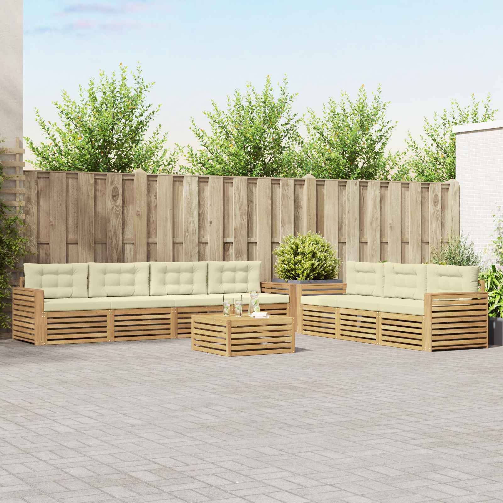 Außenterrasse mit zwei beigen Holzsofas und einem niedrigen Couchtisch auf einer gepflasterten Terrasse, vor einem Holzzaun und Grünflächen.