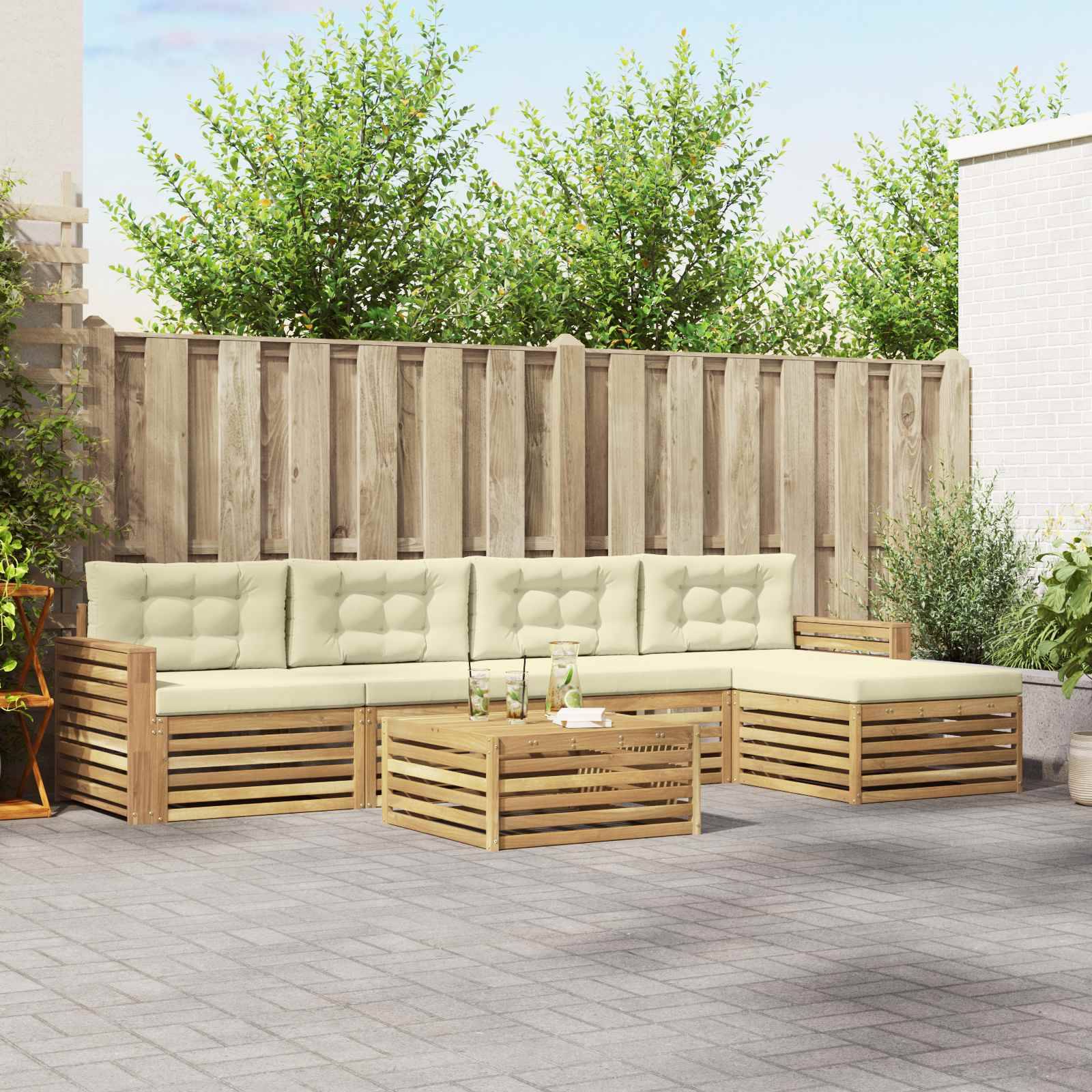 Outdoor-Terrasse mit einem Holz-Ecksofa, beige Kissen und passendem Couchtisch auf grauen Pflastersteinen vor einem Zaun und Grünflächen.