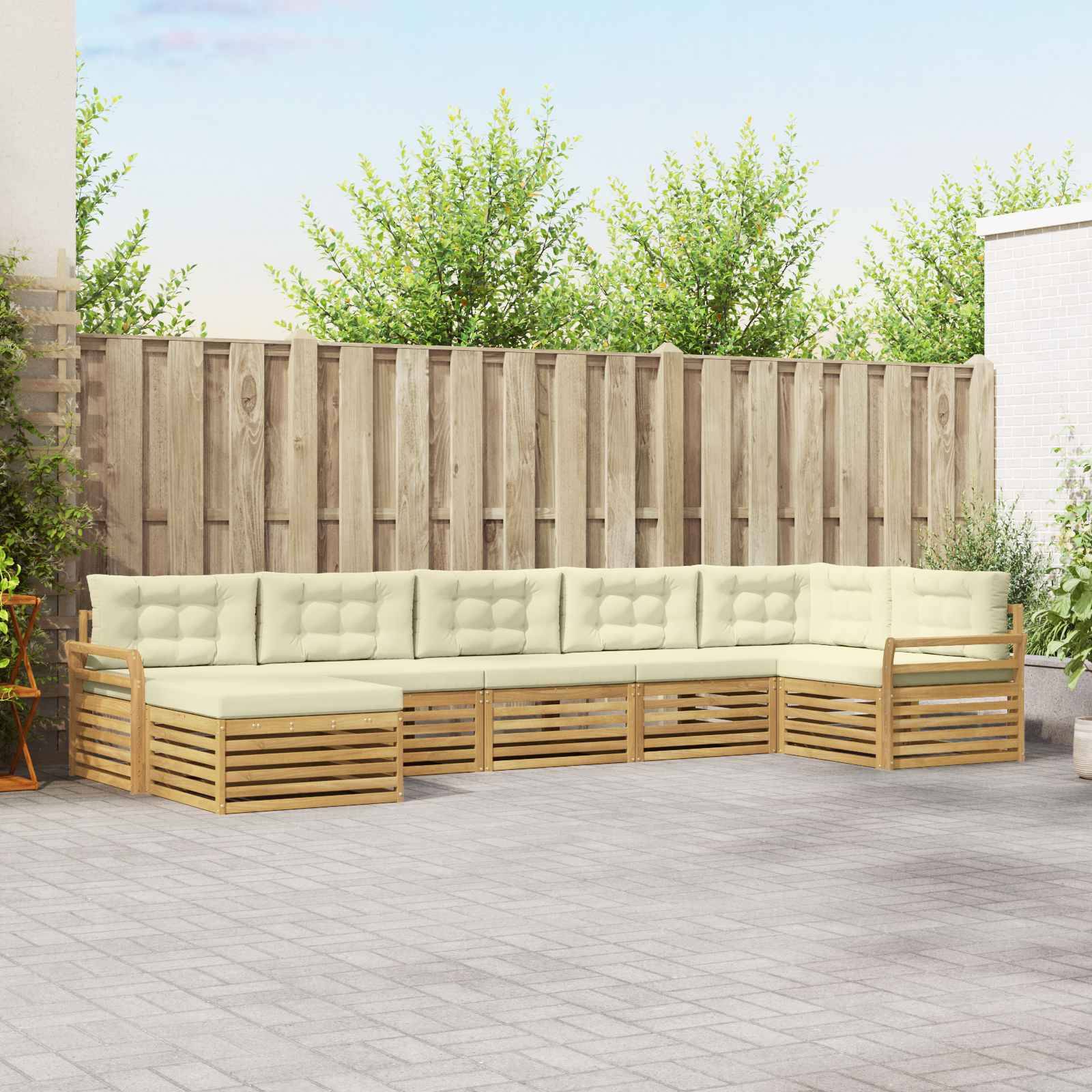 Outdoor L-förmiges Ecksofa mit cremefarbenen Kissen und Holzlattenunterbau auf einer Terrasse vor einem Holzzaun und Grünflächen.