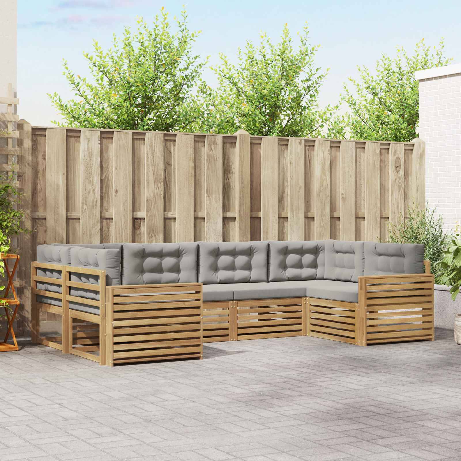 Outdoor-Sektionssofa mit grauen Kissen und Holzlattenrahmen auf einer Terrasse, vor einem hohen Zaun und Bäumen.