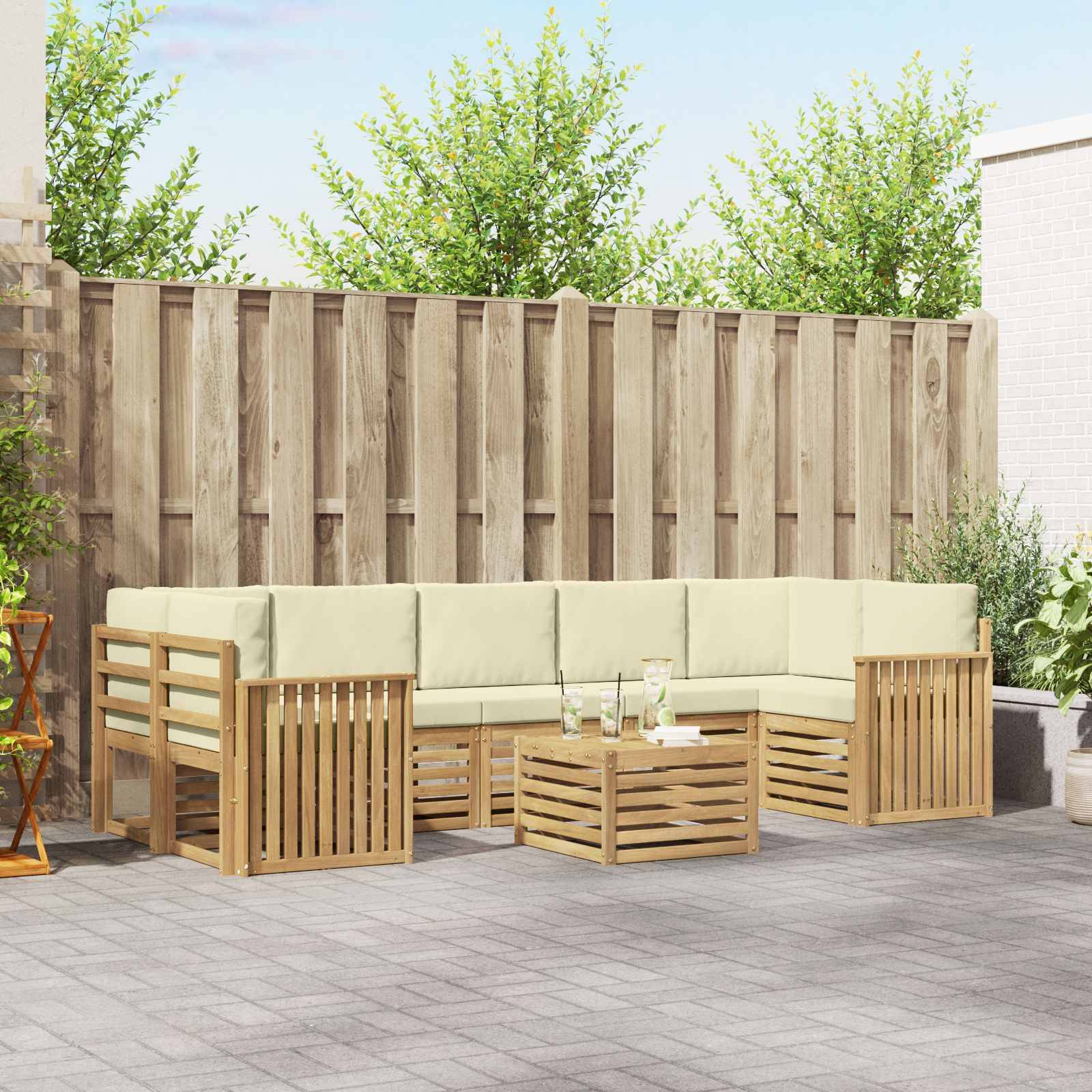 Außenbereich einer Terrasse mit einer L-förmigen Holzcouch, einem Couchtisch und leichten Kissen vor einem hohen Zaun.
