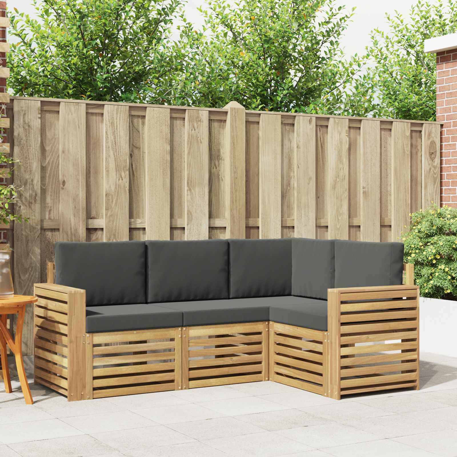 Outdoor-Ecksofa mit Holzlattenrahmen und dunklen Kissen auf einer Terrasse, vor einem Holzzaun und Gartenbegrünung.