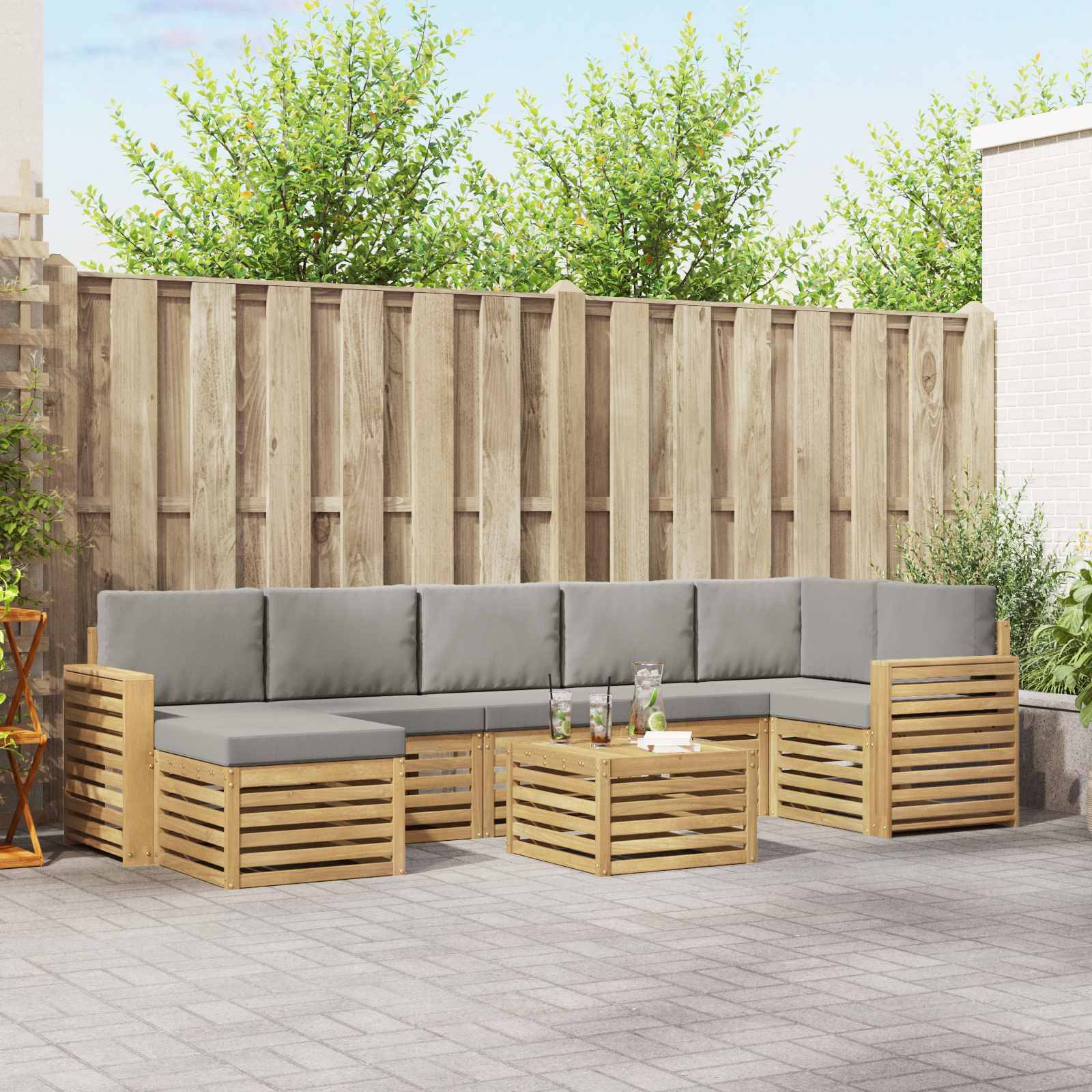 Outdoor-Terrasse mit einem L-förmigen Holz-Ecksofa und grauen Kissen um einen passenden Couchtisch, vor einem hohen Zaun und Grünflächen.