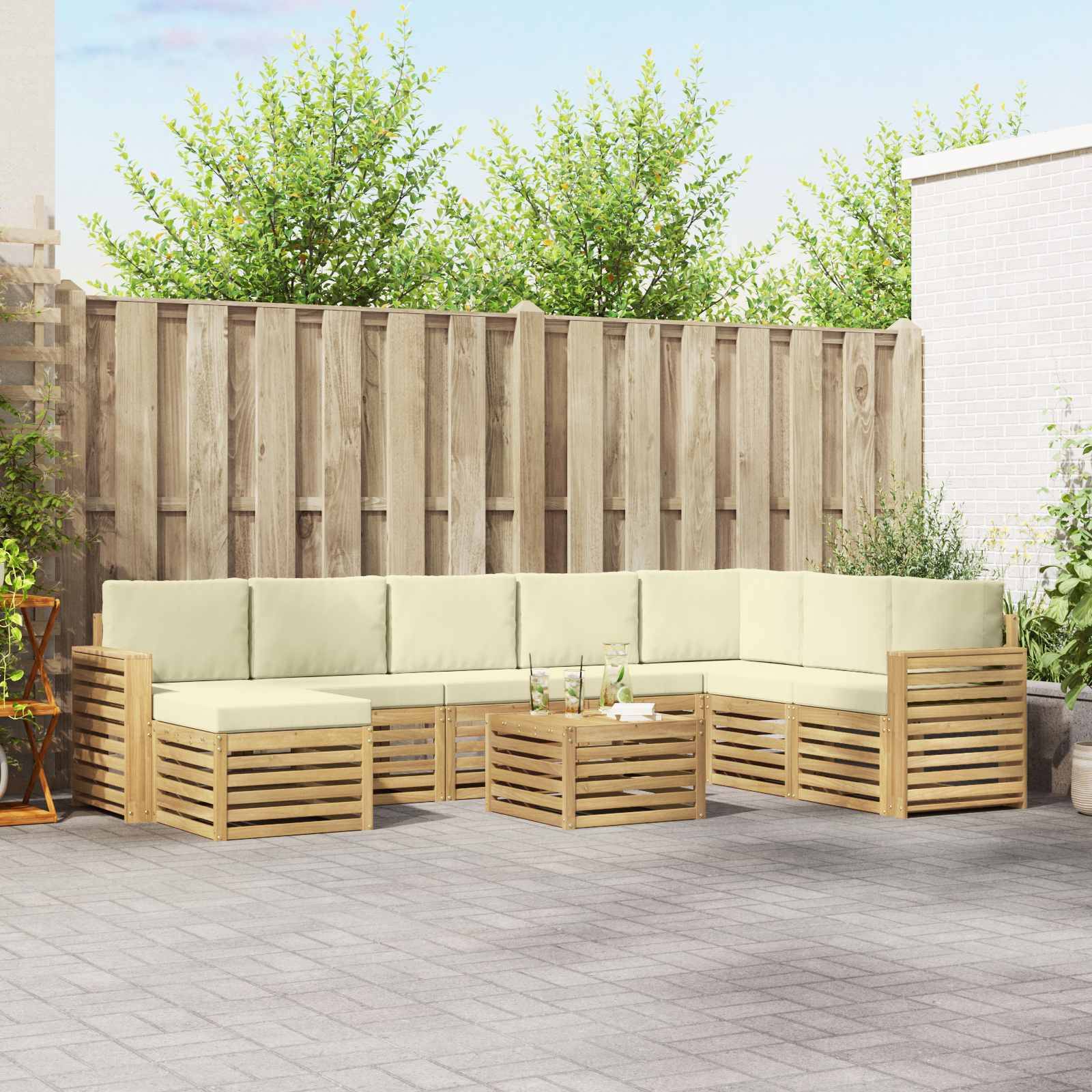 Außenterrasse mit einem L-förmigen Holzsofa und Couchtisch, beigen Kissen, aufgestellt gegen einen Zaun in einem gepflasterten Hof.