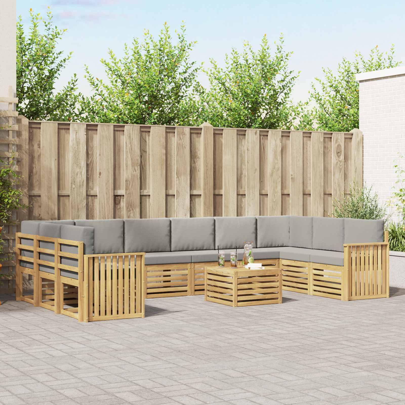 Gartenmöbel-Set für die Terrasse mit hellem Holzgestell und grauen Kissen, die um einen kleinen Couchtisch herum angeordnet sind, in einem eingezäunten Hinterhof.