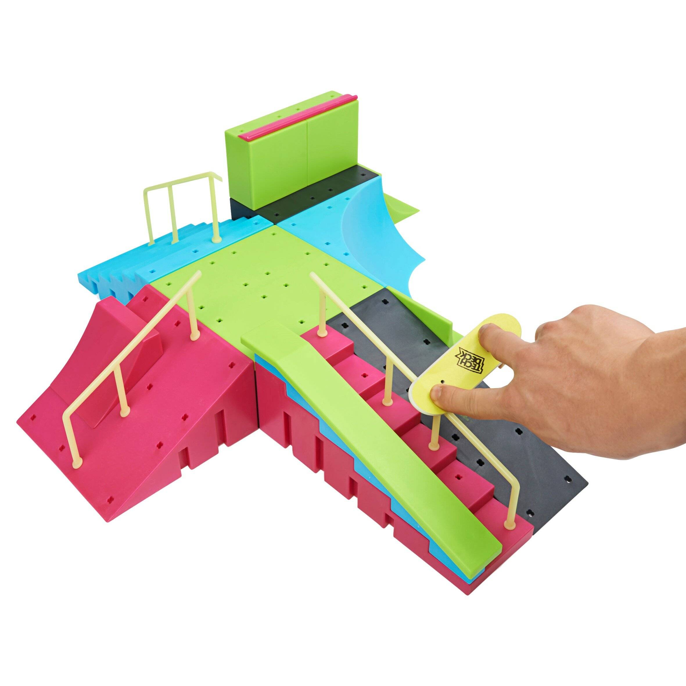 Eine Hand drückt einen kleinen gelben Knopf auf einer bunten Spielzeug-Hindernisbahn mit Rampen, Schienen und Plattformen.