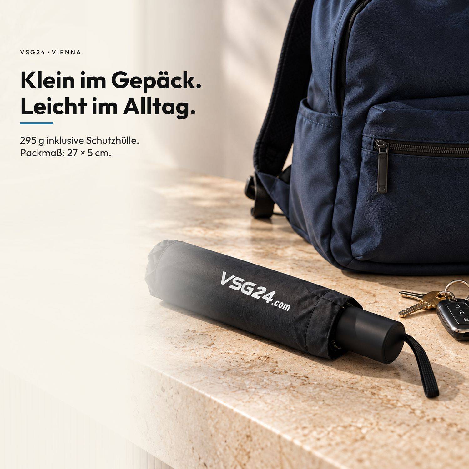 Anzeige für einen kompakten schwarzen VSG24.com-Regenschirm neben einem dunkelblauem Rucksack und Schlüsseln, mit deutschem Text: „Klein im Gepäck. Leicht im Alltag." und Spezifikationen „295 g inklusive Schutzhülle. Packmaß: 27 x 5 cm."
