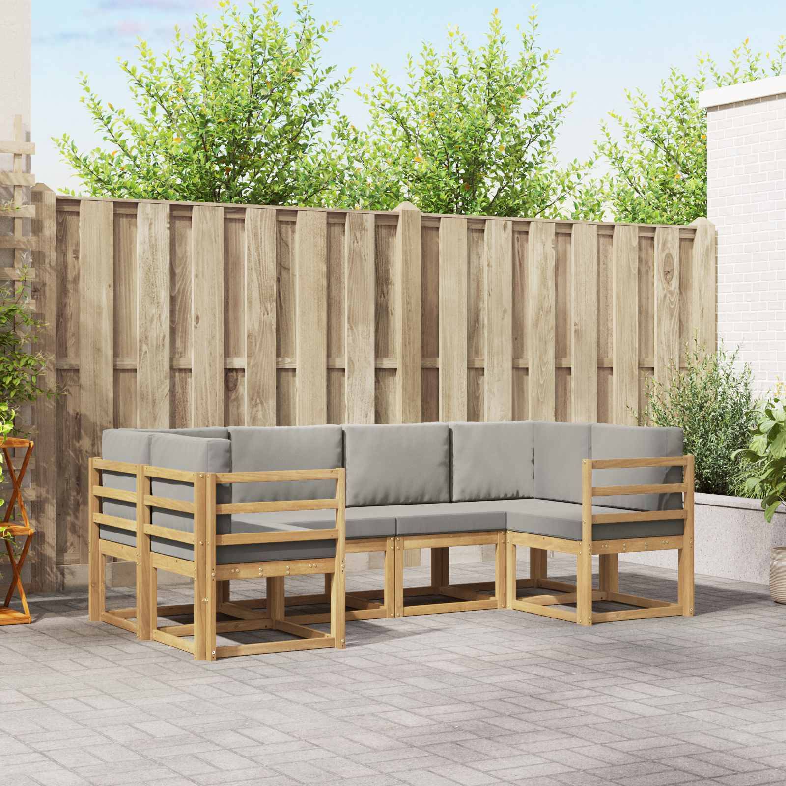 Außenpatio mit einem grauen Ecksofa mit Holzrahmen, auf einer gepflasterten Terrasse neben einem Zaun und Topfpflanzen.