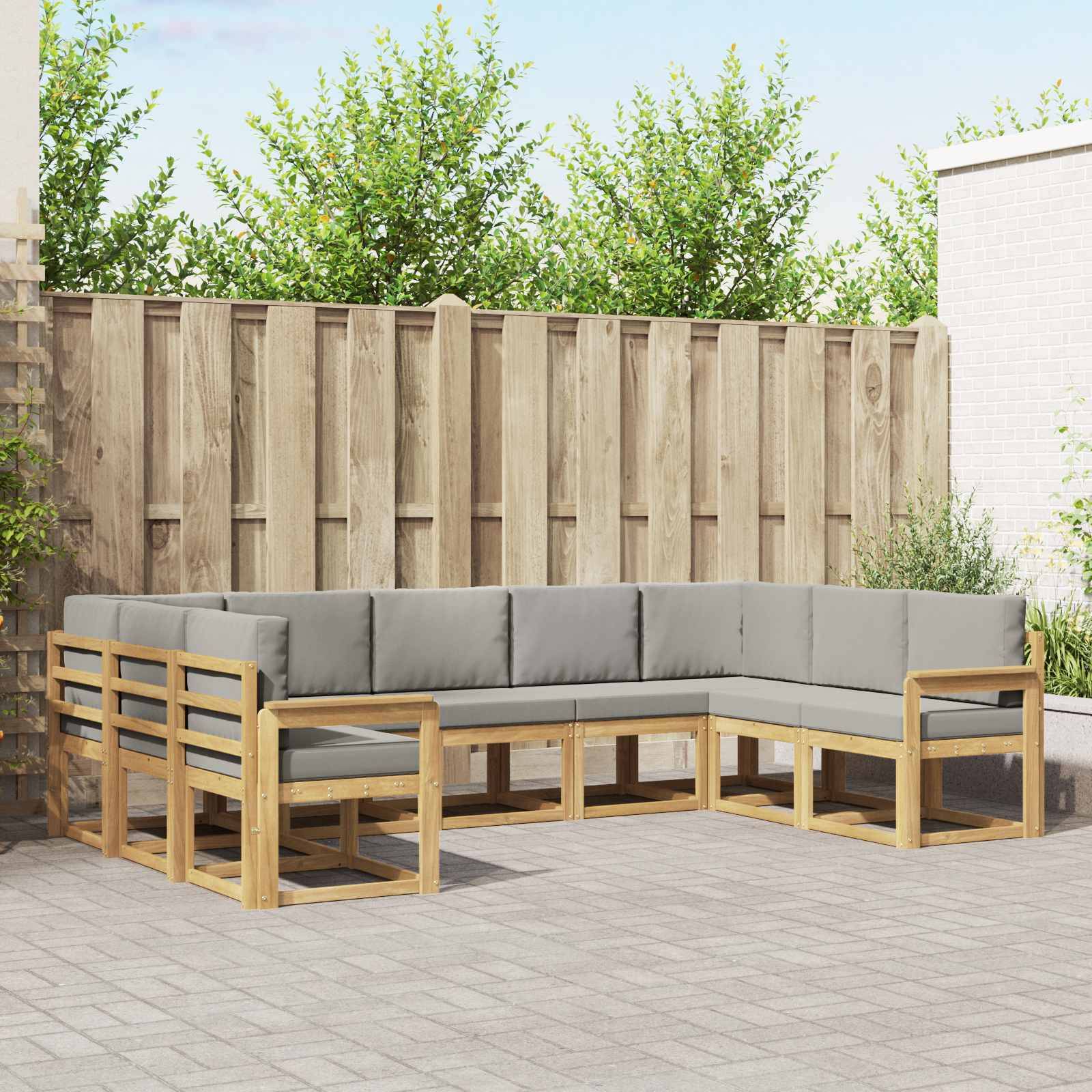 Outdoor-Sitzmöbel-Set mit hellem Holzrahmen und grauen Kissen, angeordnet in einer L-förmigen Sektionalgarnitur um einen niedrigen Tisch, vor einem Holzzaun.