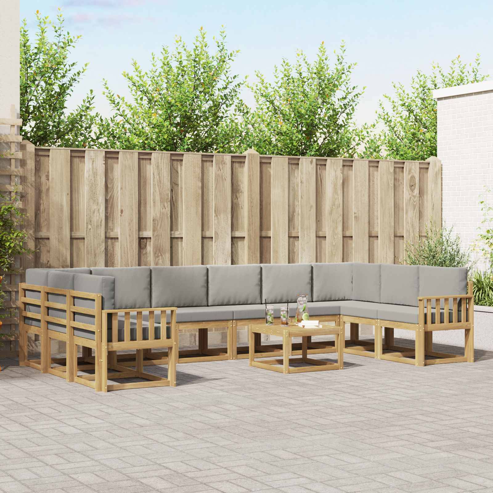 Außenterrasse mit einem hellen Holz-Ecksofa, passenden Sesseln und Couchtisch vor einem Holzzaun und Grünflächen.