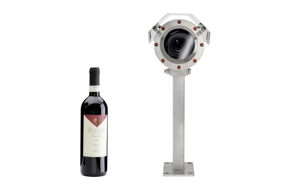 Flasche Rotwein neben einer metallischen Industriekamera oder einem Objektiv auf einem Stativ vor weißem Hintergrund.