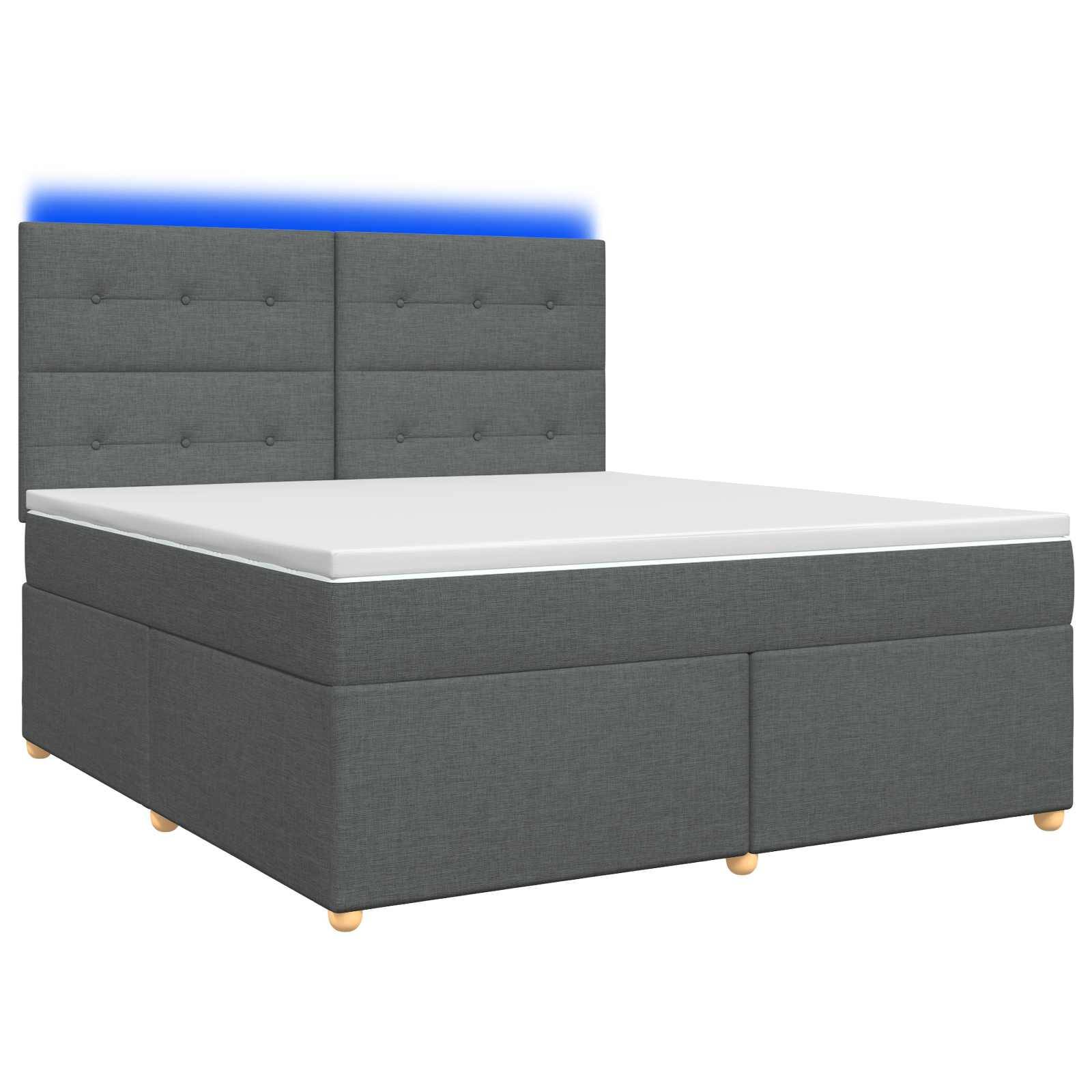 Grau gepolstertes Bett mit gestepptem Kopfteil, weiße Matratze und niedrige Holzfüße auf neutralem Hintergrund.