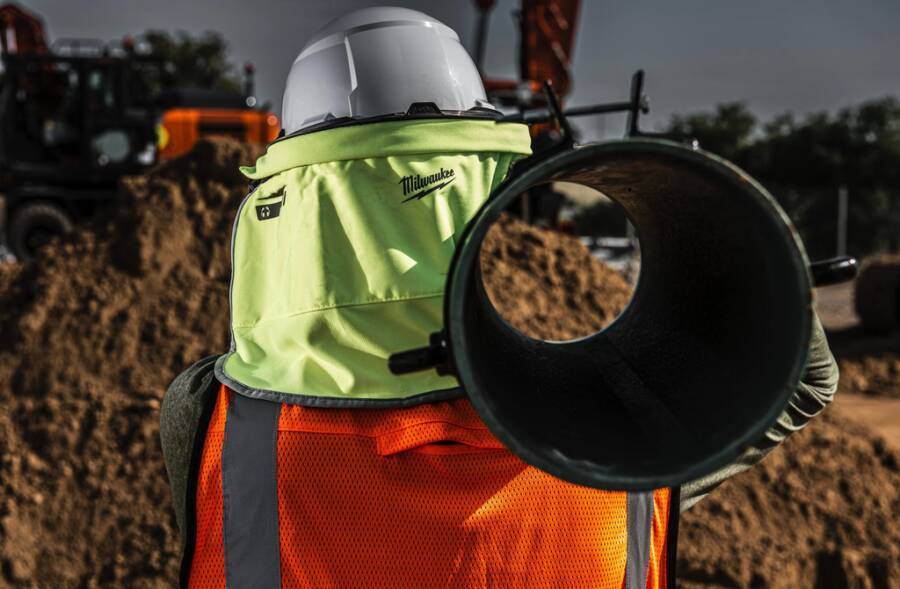 Bauarbeiter in Schutzhelm und Warnweste hält ein großes Rohr oder Rohrpaar auf einer Baustelle mit Maschinen im Hintergrund.