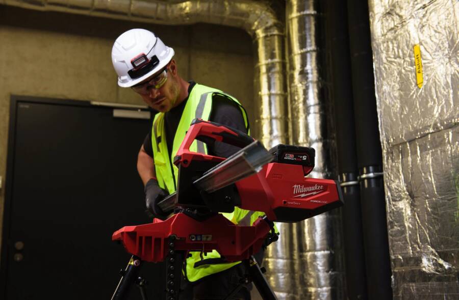 Bauarbeiter in Schutzhelm und Sicherheitsweste, der ein rotes Milwaukee-Werkzeug an einem Metallrohr in einem industriellen Technikraum einsetzt.