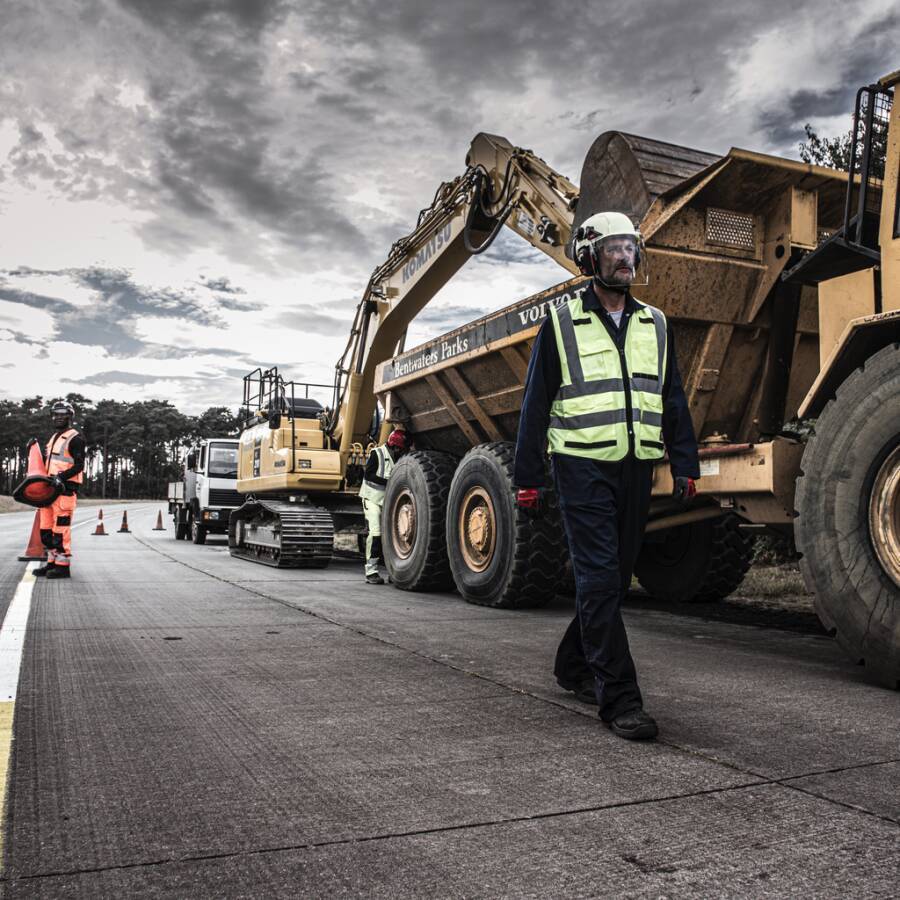 Straßenarbeiter in Warnkleidung stehen neben einem Kipplaster und einem Bagger auf einer Autobahnbaustelle unter bewölktem Himmel.