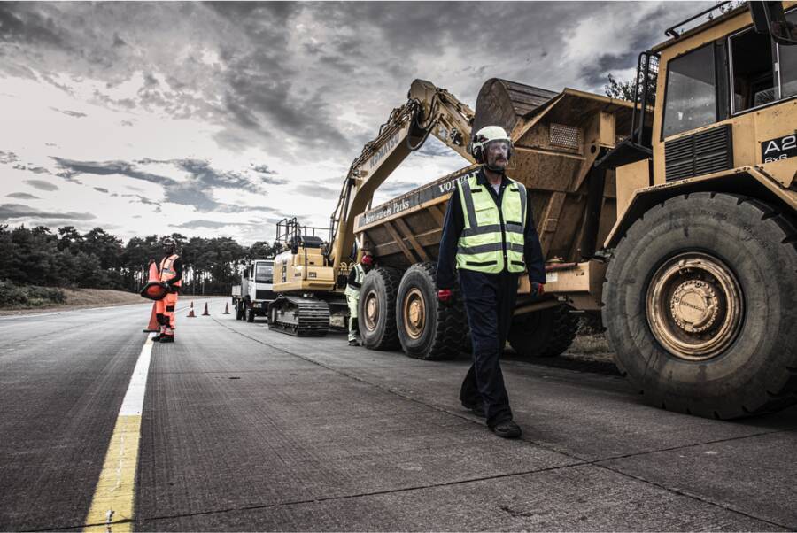 Bauarbeiter und schwere Straßenbaumaschinen auf einer Baustelle zur Straßenerneuerung an einer Autobahn, mit einem Kipper, einem Bagger und Verkehrskegeln, die das Arbeitsgebiet kennzeichnen.