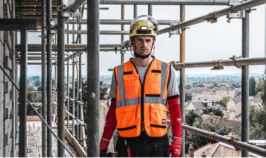 Bauarbeiter in orangefarbener Sicherheitsweste und Helm, der auf Baugerüsten auf einer Baustelle steht, mit Stadtblick hinter ihm.