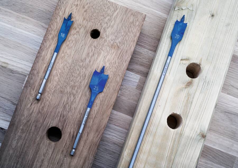 Drei flache Holzbretter mit gebohrten Löchern und eingefügten blauen Spatenbohrbits, die unterschiedliche Lochgrößen in einem Holzbearbeitungsvergleich zeigen.