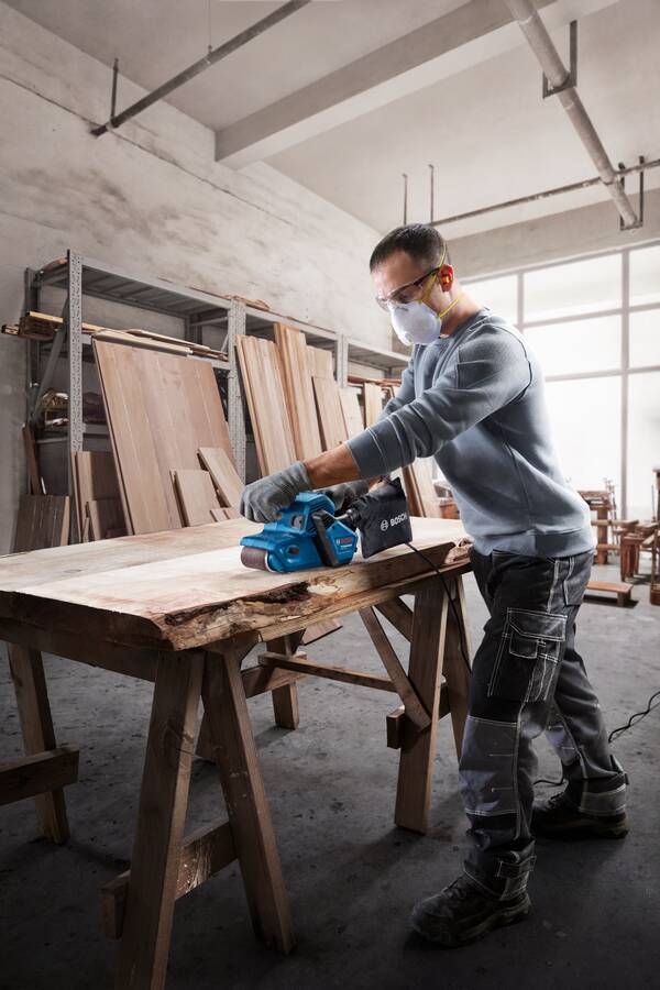Schreiner in einer Werkstatt trägt eine Maske und Handschuhe und schleifen ein großes Holzbrett mit einem Elektroschleifen auf Böcken.