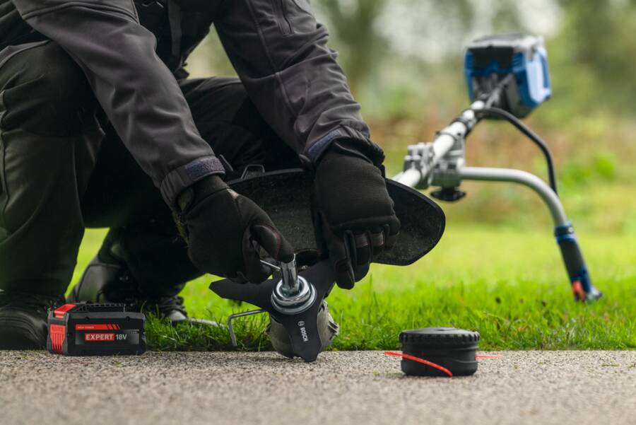 Person in Handschuhen, die einen tragbaren Fadentrimmer verwendet, um Gras entlang eines Gehwegs zu kanten, mit einem Akku und einem Werkzeug auf dem Boden.