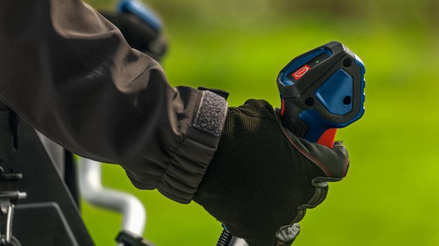 Behandschuhte Hand, die einen blau-roten Joystick-Controller an einer Maschine greift, wahrscheinlich zum Bedienen eines Fahrzeugs oder einer Ausrüstung.
