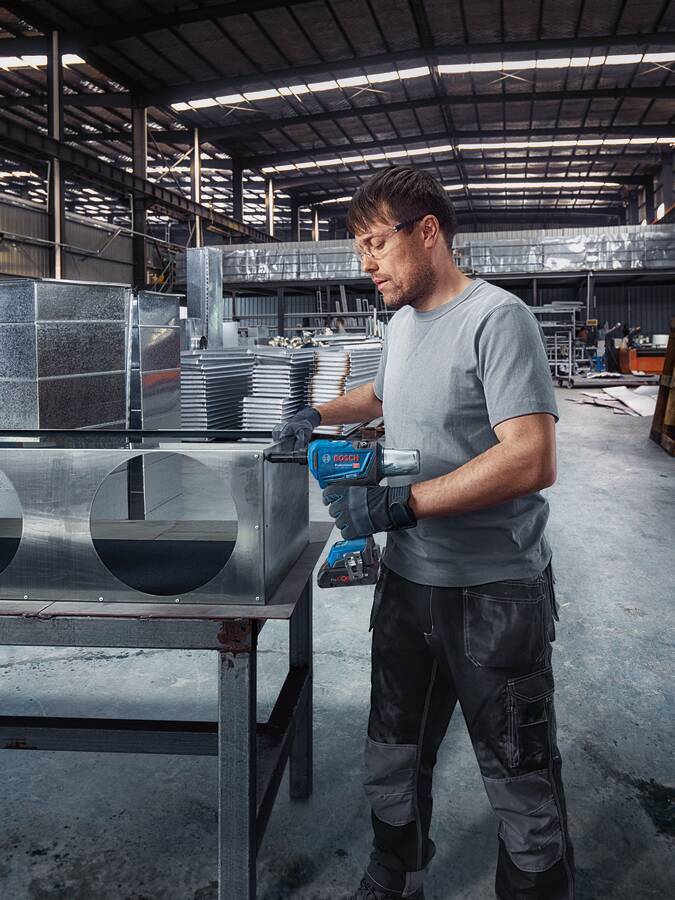 Arbeiter verwendet einen Elektroschrauber an einer großen Metallkiste in einer Industriewerkstatt, mit Stapeln von Blechen im Hintergrund.