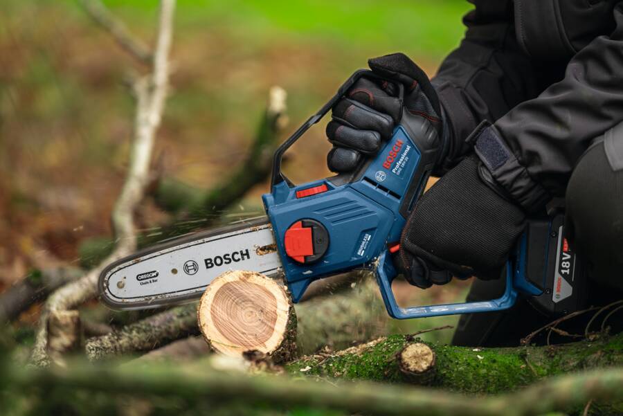 Eine handschuhtragende Person nutzt eine Bosch-Akku-Kettensäge, um einen gefallenen Baumstamm im Freien in einem bewaldeten Gebiet zu schneiden.