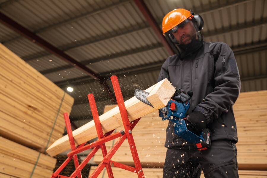 Arbeiter mit Schutzhelm und Sicherheitsausrüstung, der in einer Werkstatt einen Holzbalken mit einer Kettensäge zuschneidet, wobei Holzspäne umherfliegen.