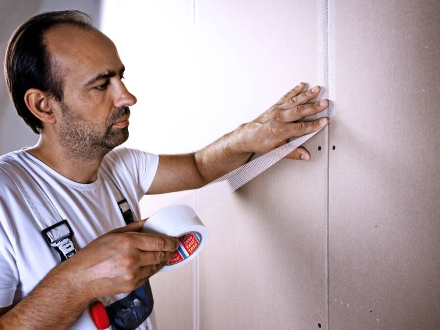 Ein Mann bringt Trockenbandaufkleber auf einer Wandfuge an, hält eine Rolle Klebeband, während er es auf die Fuge drückt.