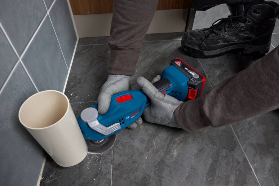 Person, die einen kabellosen Winkelschleifers verwendet, um eine Bodenfliese neben einer Wand zu schneiden oder zu schleifen, mit einem Staubsammelbehälter in der Nähe.