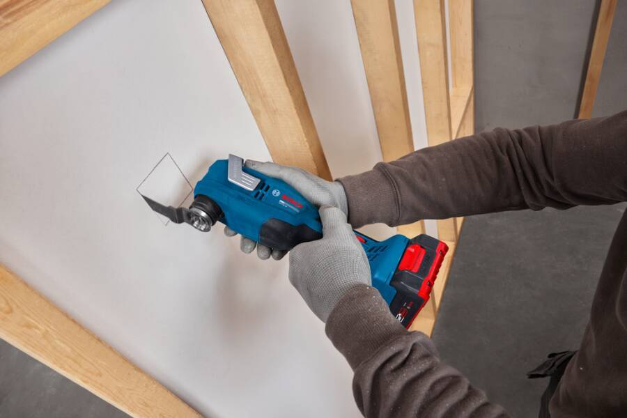 Eine Person, die mit einem schnurlosen Elektrowerkzeug ein weißes Wandpaneel in einem Raum mit Holzständern schneidet oder bohrt.