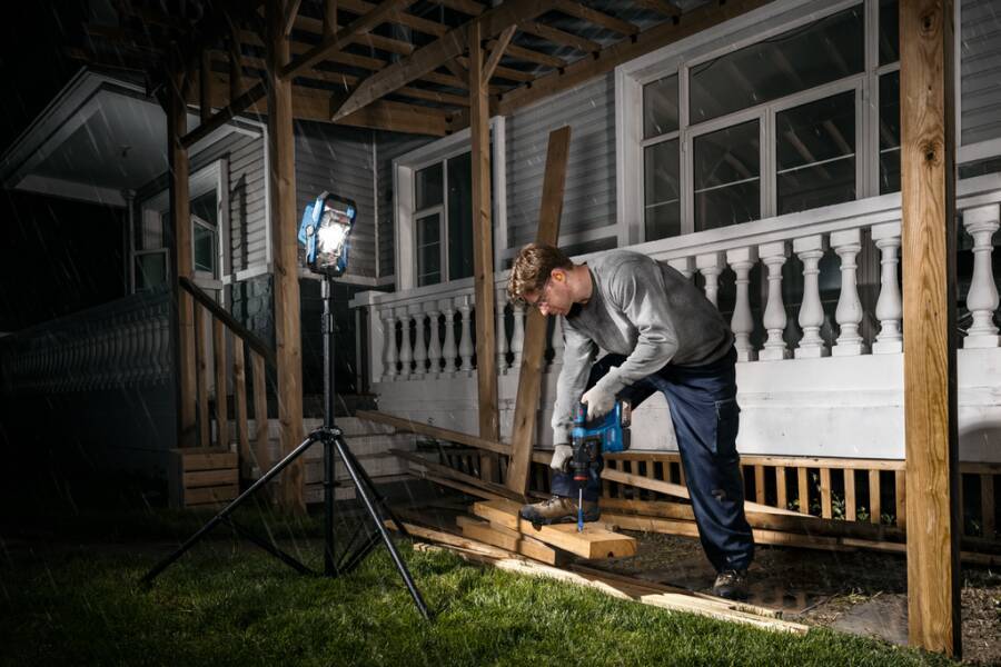 Ein Arbeiter bohrt nachts in einen hölzernen Pfosten auf einer Veranda, wobei er eine Dreibein-Arbeitsleuchte und eine Elektrobohrmaschine unter der Hausfassade verwendet.