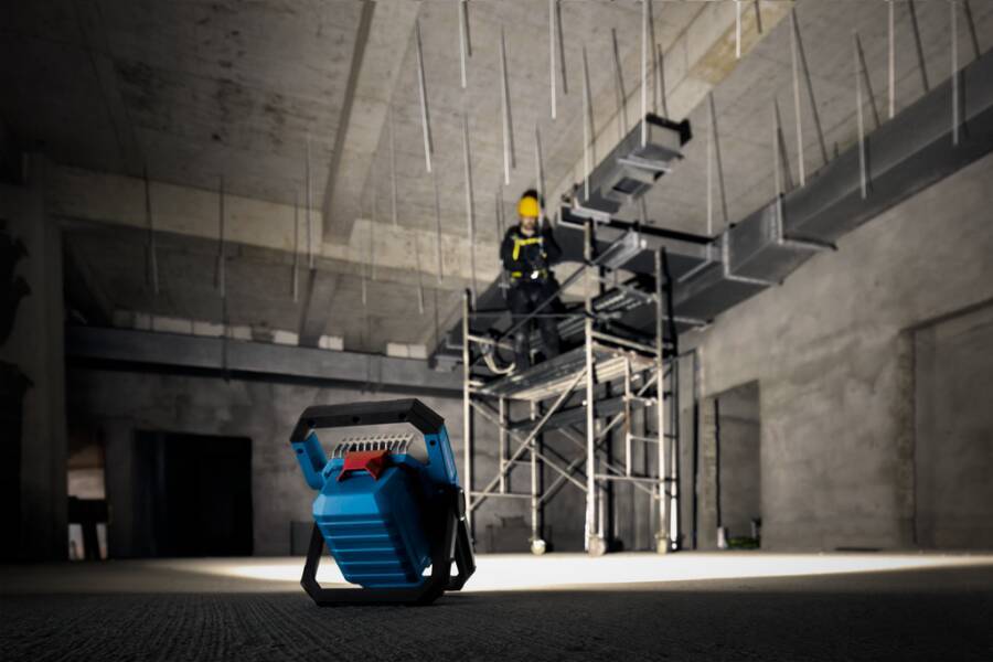 Arbeiter in Schutzausrüstung auf einem Gerüst inspiziert ein unfertiges Betoninneres, während ein blaues tragbares Arbeitslicht die dunkle Baustelle beleuchtet.