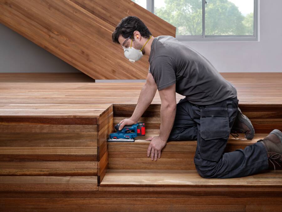 Maskierter Handwerker kniet auf hölzernen Treppen und schleifen oder versiegelt eine Stufe mit einem tragbaren Elektrowerkzeug in einem modernen Haus.