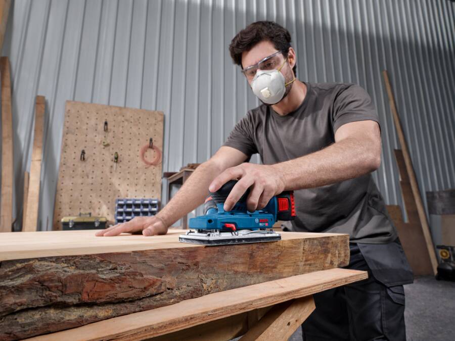 Ein Mann, der eine Schutzbrille und eine Staubmaske trägt, schleiht ein Holzbrett mit einem Elektroschleifera in einer Werkstatt.