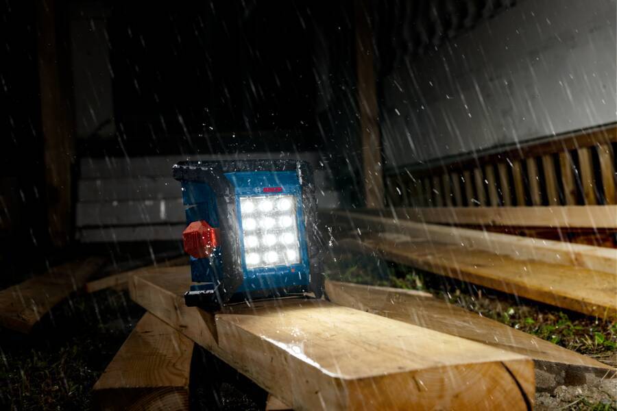 Tragbare LED-Arbeitsleuchte, die nachts bei starkem Regen auf Holzbretter scheint, wahrscheinlich im Freien auf einer Baustelle.