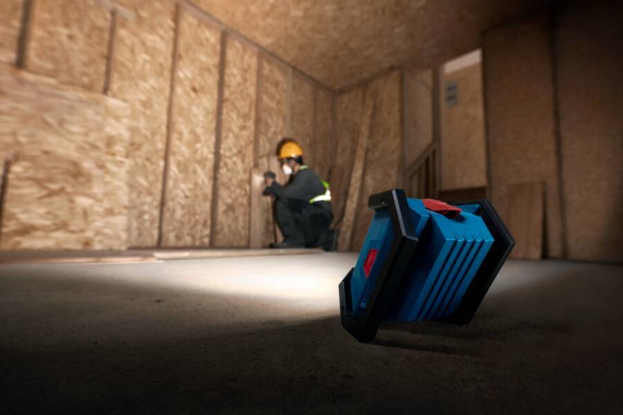 Arbeiter in einem Schutzhelm kniet in einem Sperrholzzimmer, während ein blaues tragbares Gerät im Vordergrund auf dem Boden steht.