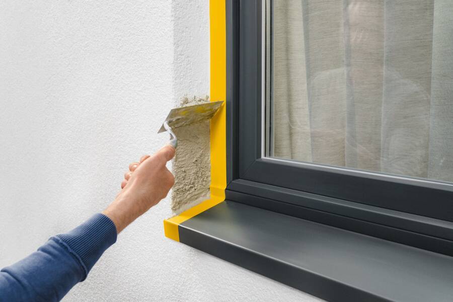 Person, die Gips oder Dichtungsmittel auf die Wandkante neben einem Fenster aufträgt, während Abdeckband den dunklen Fensterrahmen schützt.