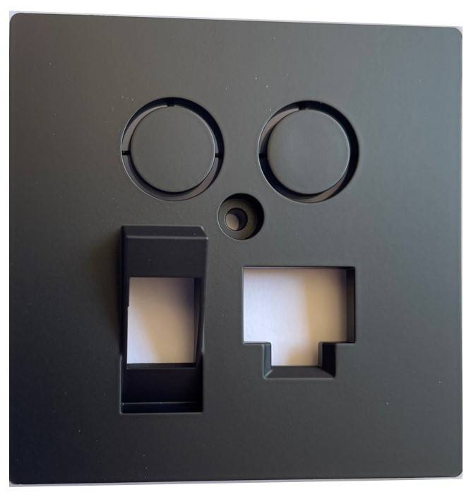 Schwarze quadratische Frontplatte mit zwei kreisförmigen Öffnungen, einem kleinen Mittelloch und zwei rechteckigen Ausschnitten, wahrscheinlich eine Wandsteckdose oder Schalterabdeckung.