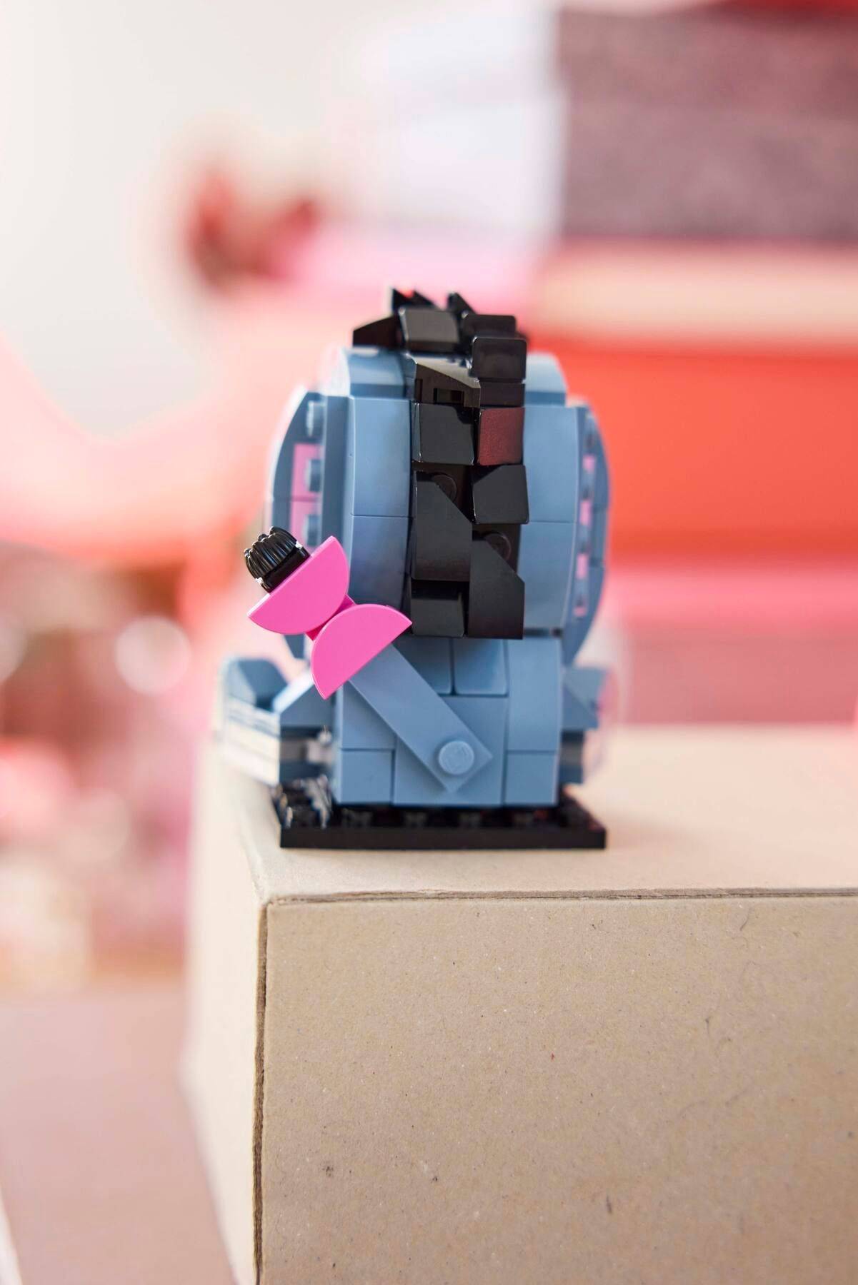 Kleine LEGO-ähnliche blaue Roboter- oder Spielzeugfigur mit schwarzem Gesichtspanel und rosa Schleife, die auf einem Karton in Innenräumen sitzt.