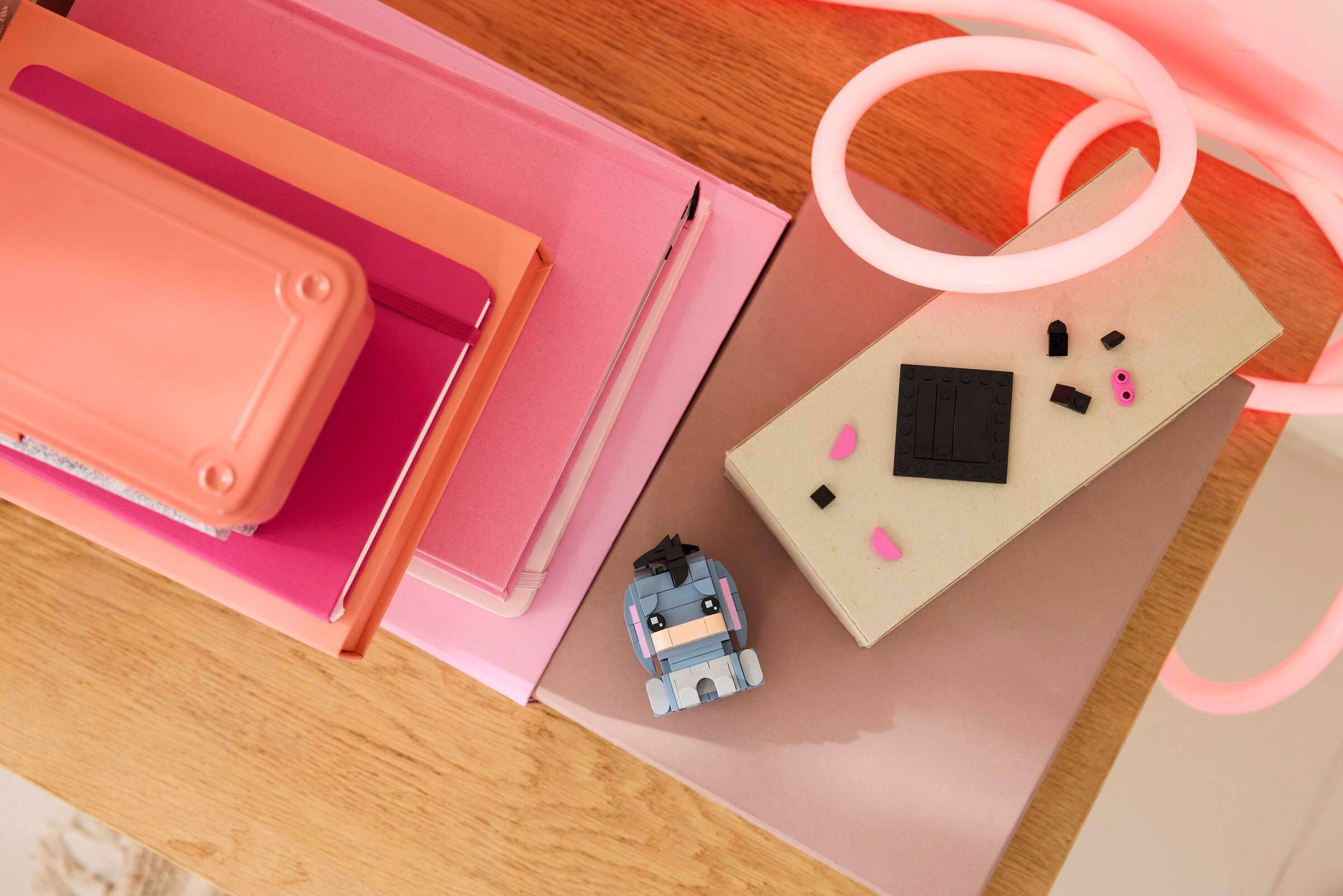 Draufsicht auf einen rosa und beigefarbenen Arbeitsplatz mit gestapelten Notizbüchern, einem Stifteetui, einer kleinen Spielfigur und einer Schreibtischlampen-Schlinge auf einem Holztisch.