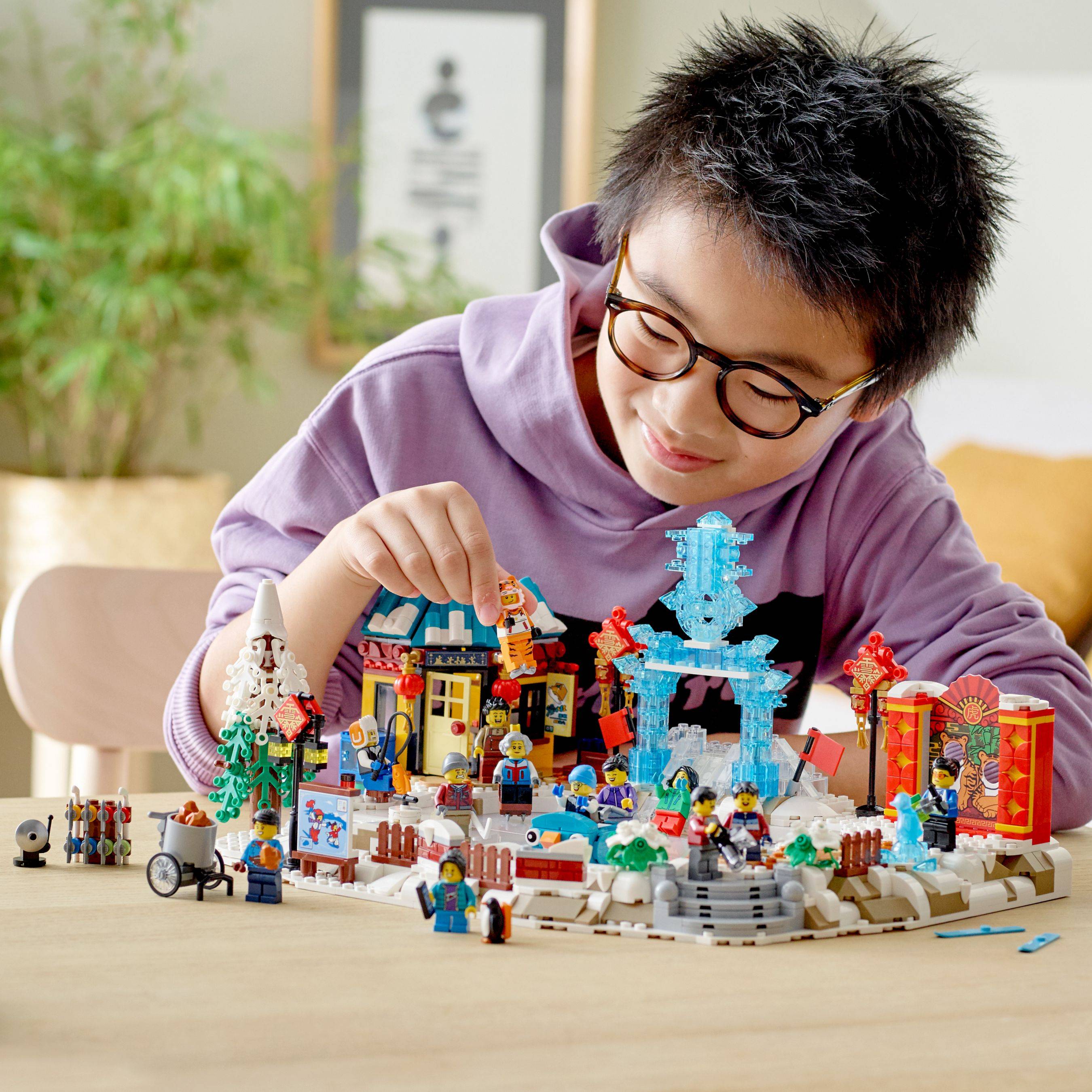 Ein Kind mit Brille und violettem Hoodie spielt mit einer bunten LEGO-ähnlichen Miniaturstadt-Szene auf einem Tisch.